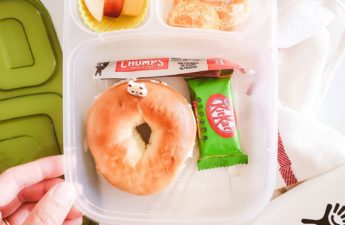 a hand touching a school lunch box with bagel, green tea kitkat, beef jerky, apples and oranges